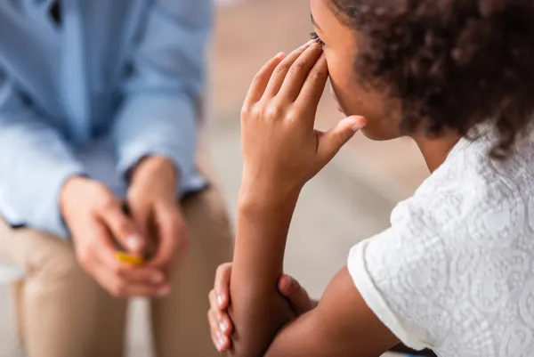 Close up view of girl with hand near eye crying during consultation with blurred counsellor on background